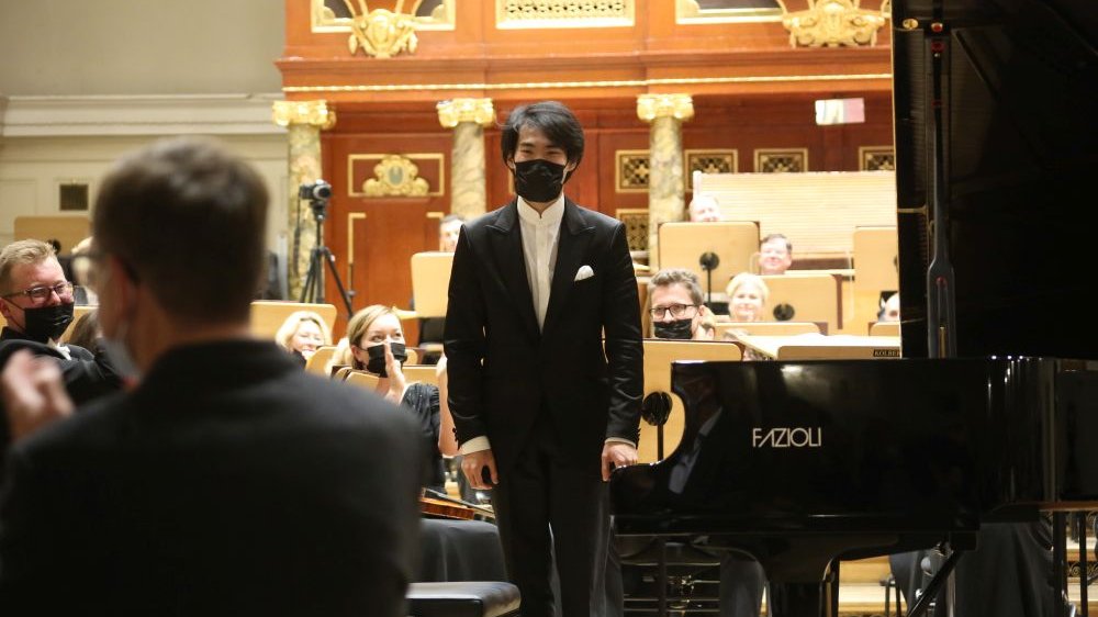 Picture from the concert: Bruce Liu standing next to the grand piano. THe members of the orchestra in the background.