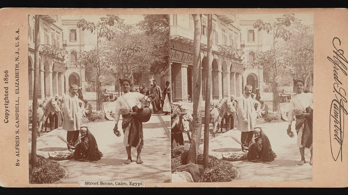 Stereophotograph of an outdoor square in sepia colours: three people in a foreground - one carrying something resembling a clay vessel, one sitting on the ground and one standing next to him or her. Other people, a horse and a building with arcades as a background.