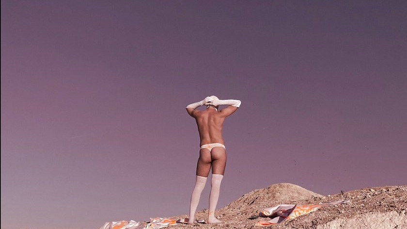 One of the works from the exhibition: a picture of half-naked man standing on a rock, wearing white knee socks, long white gloves, white cap and white thong.