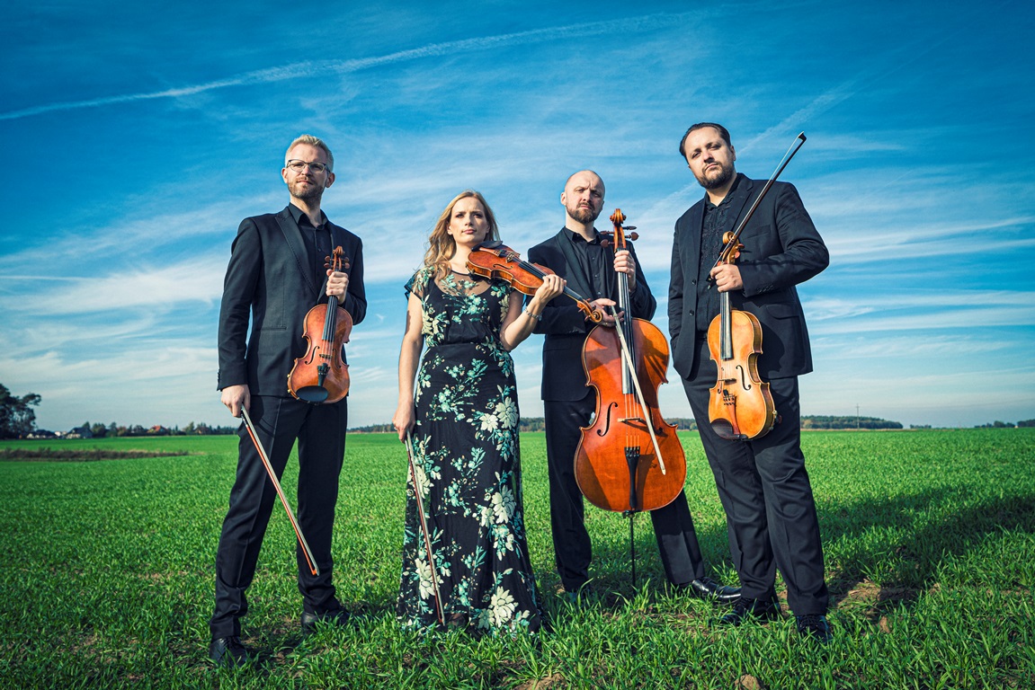Photo of the Meccore String Quartet members: one woman and three men holding their musica instruments and standing on the grass. Blue sky as the background. - grafika artykułu