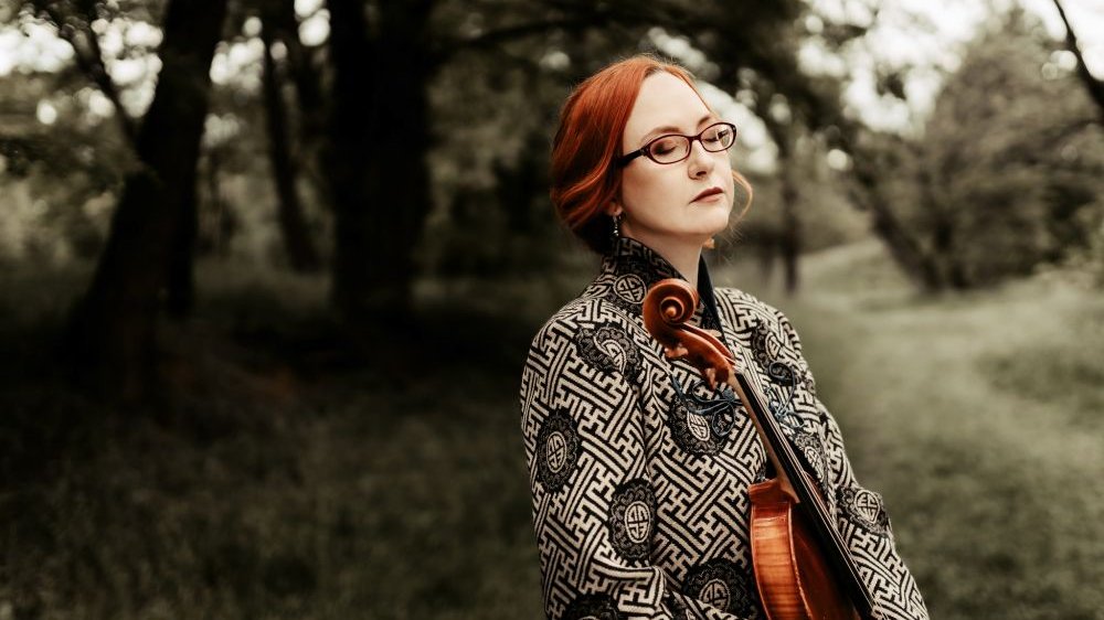 Photo of the artist - a woman with closed eyes holding the violin; trees as the background. - grafika artykułu