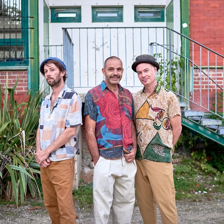 Photo of the band: three men in colourful shirts; as a background - a part of a building with stairs and some plants. - grafika artykułu