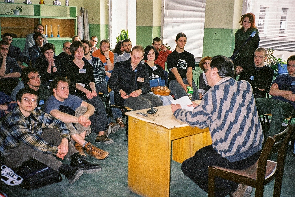 A group of people sitting and listening to the lecture in a classroom. In the foreground a man sitting at the desk, holding papers in his hands. - grafika artykułu