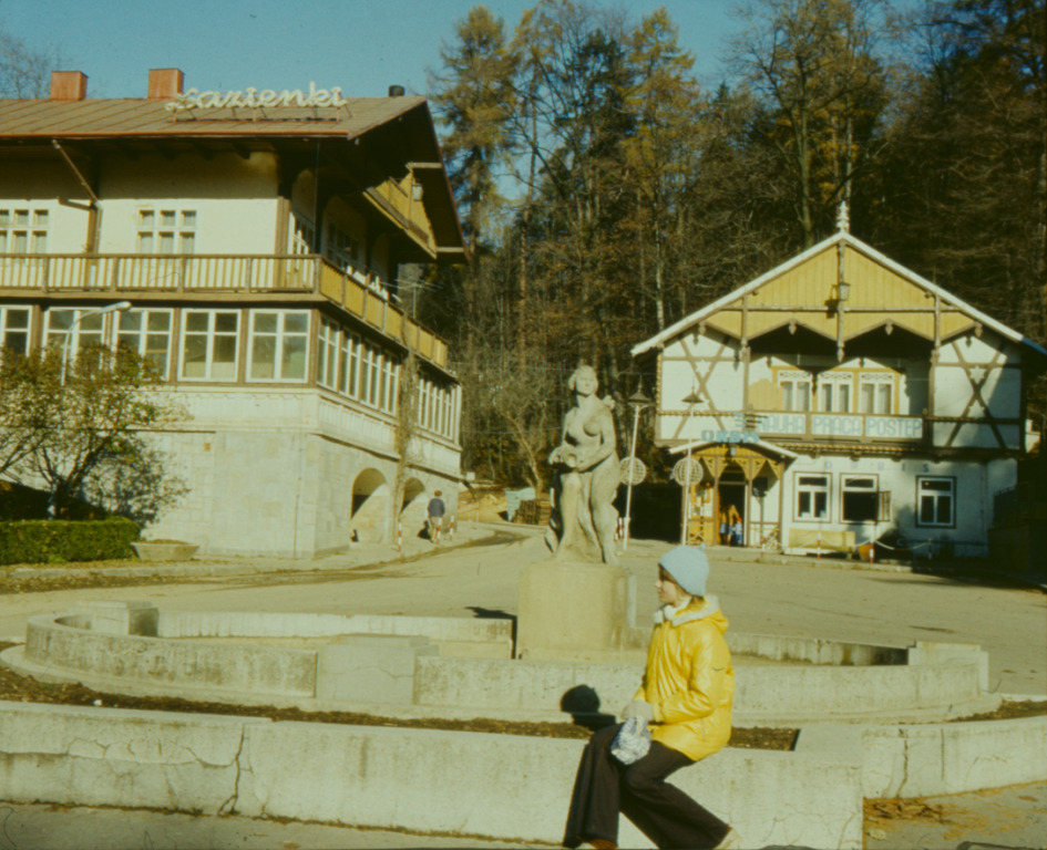 Photo of a girl sitting on a concrete parapet in front of a sculpture. Two buildings in a background. - grafika artykułu