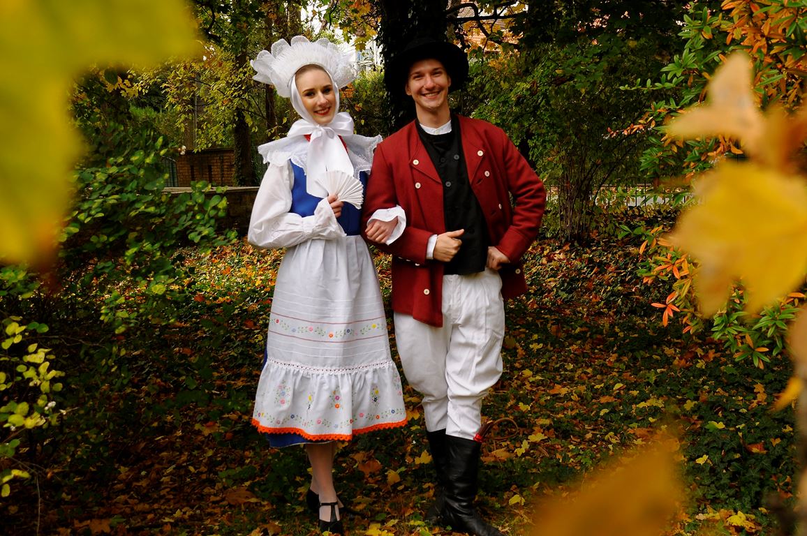 Photo of a woman and a man dressed in traditional folk costumes. Trees and colourful leaves as a background. - grafika artykułu
