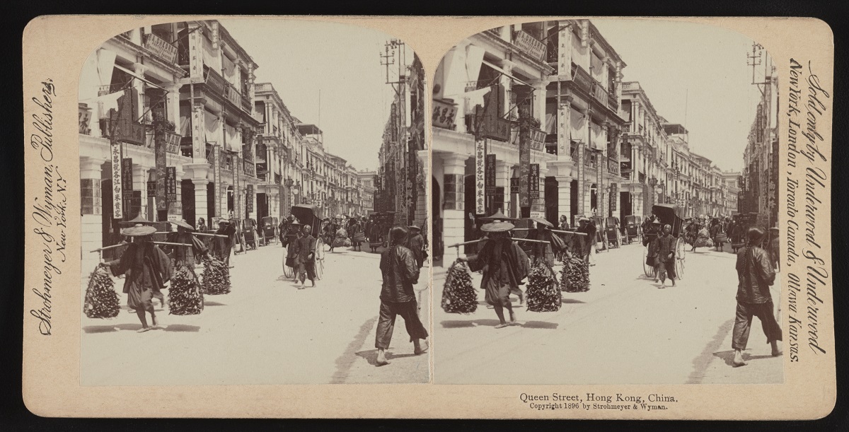 Stereophotograph of a street in sepia colours: in the foreground two people carrying goods on their shoulders. Lane of buildings on the left. - grafika artykułu