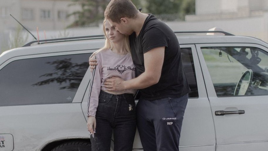 Photo of a couple standing by a car. The young man is hugging a girl.