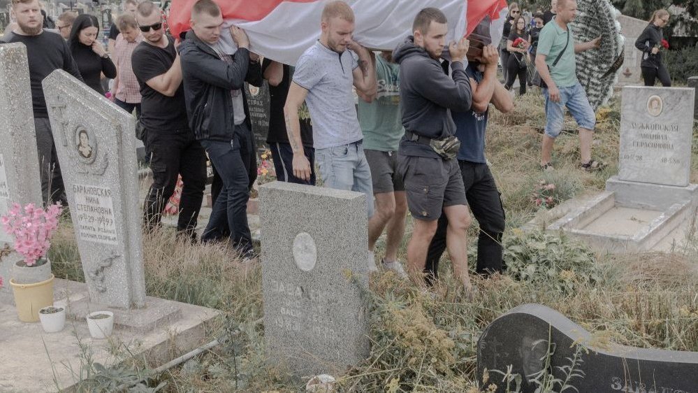 Photo of men carrying a coffin covered with a historic belarusian flag. The men and other people go between graves on the cemetery.