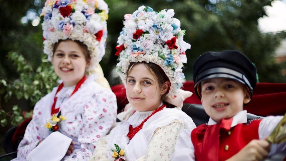 Picture of two girls and a boy in traditional national costumes. The girls have decorative headgears on their heads.