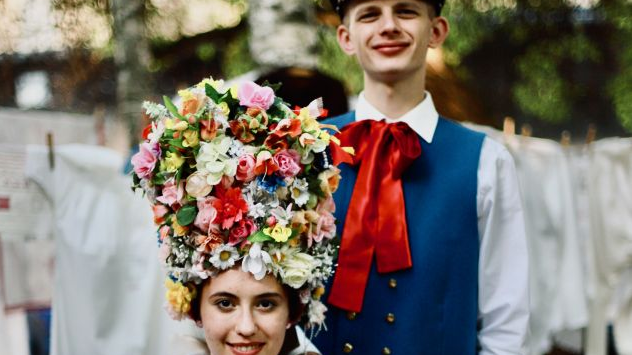 Young girl and a young man in traditional costumes. The girl is sitting, the man is standing behind her. The girl has decorative headgear on her head.
