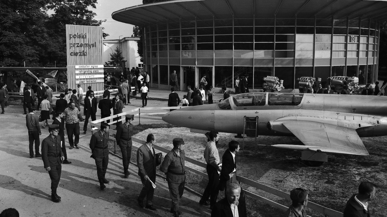Black and white photo of a round building - a fair pavilion. In the foreground - a military plane and people walking around and looking at it.