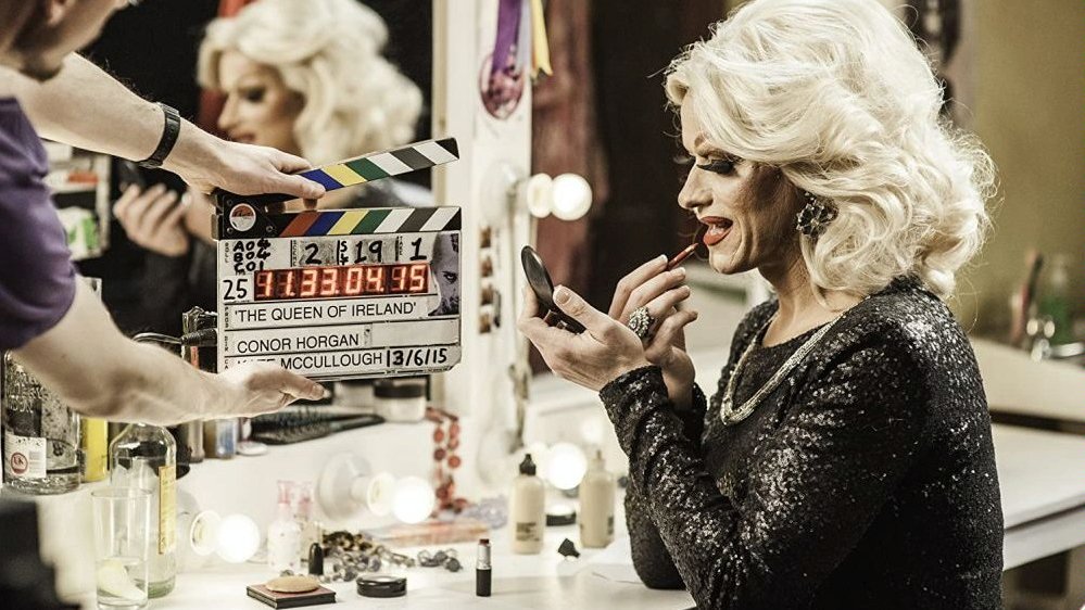 A blond woman in black dress who is sitting by the dressing table and putting her lipstick on. On the left - a man holding a clapperboard.