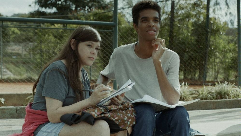 Photo from the movie: two teenagers (a girl and a boy) sitting outside and looking at something. A girl is holding a notebook and a ballpen. A fence and some trees in the background.