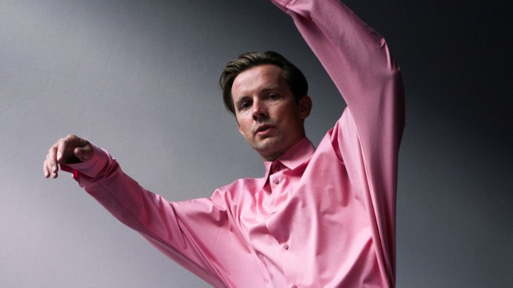 Młody mężczyzna w różowej koszuli unosi ręce do góry na tle szarej ściany.