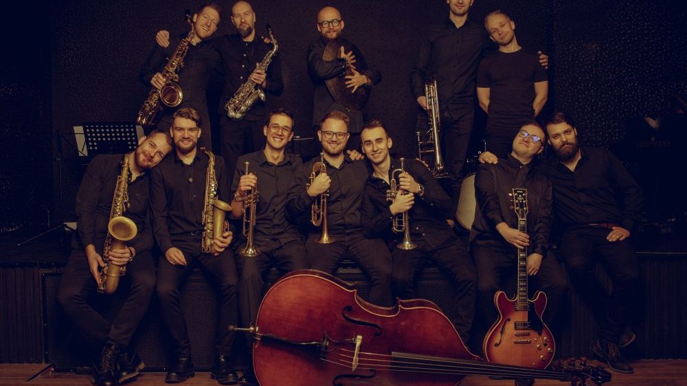 Dwunastu mężczyzn ubranych na czarno siedzi obok siebie, trzymają swoje instrumenty, które lśnią na złoto - trąbki, saksofony, puzony czy gitary.