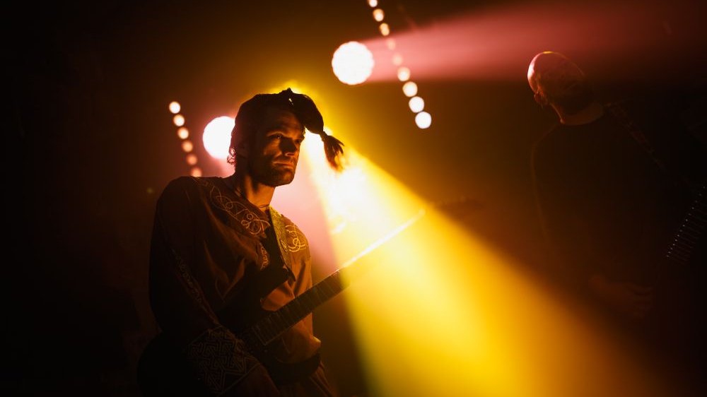 Mężczyzna w ludowej koszuli i kitce na głowie stoi na scenie. Pod pachą trzyma gitarę elektryczną, z tyłu pada żółte światło.