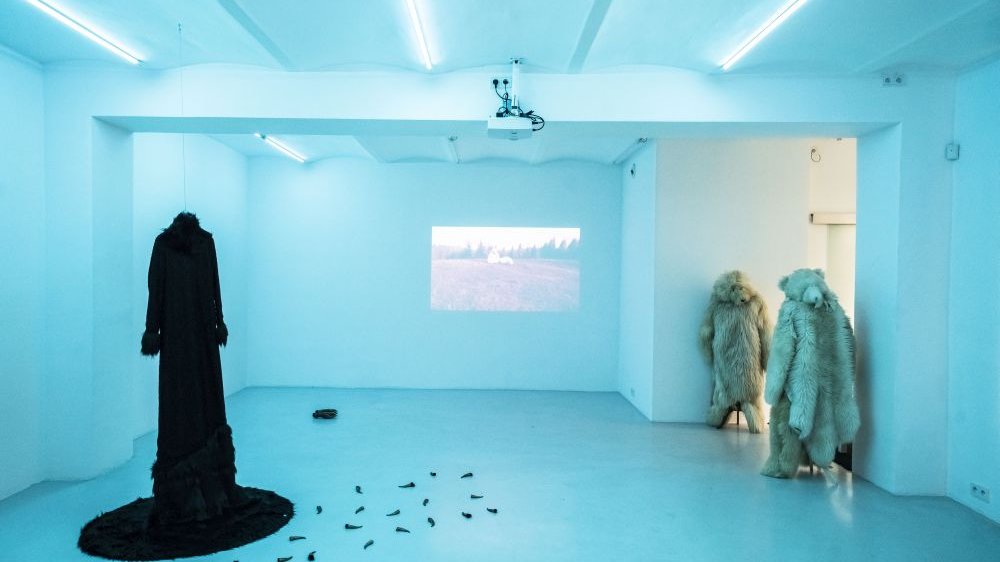 Jasna, oświetlona sala wystawowa z czarną suknią, dwoma kostiumami niedźwiedzia, rozsypanymi na podłodze pazurami i projekcją video na ścianie.