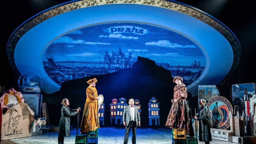Na scenie widać wielki obraz miasta Praga, podpisany "Praga". Na samej scenie stoi kilku mężczyzn - dwóch z nich na podestach, w bogatych strojach i czepcach.