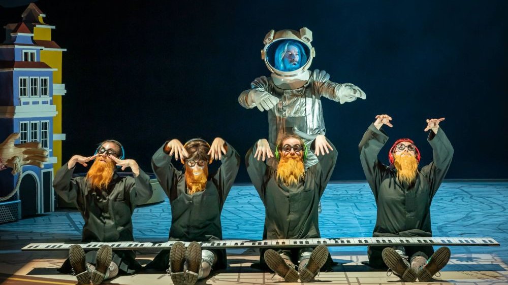 Bohater w stoju kosmonauty przechadza się wokół czwórki tak samo ubranych postaci, każda z nich ma okrągłe okulary i płomiennorudą brodę.