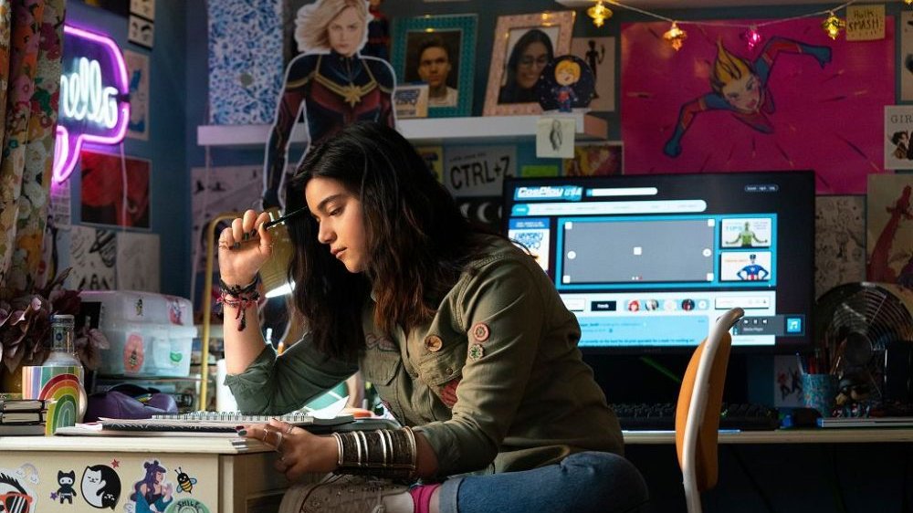 Dziewczyna siedzi przy biurku, podpiera głowę ołówkiem. Za nią komputer i ściana oklejona plakatami.