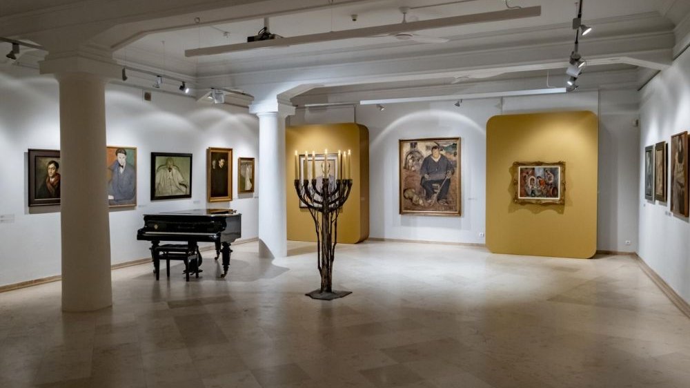 Sala muzeum z obrazami na ścianach, fortepianem po lewej i menorą z palącymi się świecami na środku.