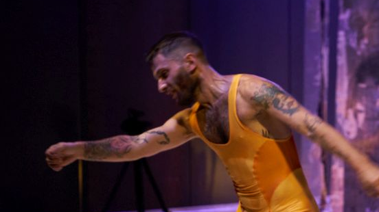 Mężczyzna w żółtym, obcisłym kombinezonie tańczy na scenie.