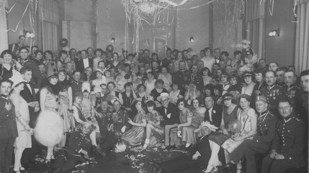 Tłum ludzi na balu pozuje do zdjęcia grupowego. Nad ich głowami wielki żyrandol i ozdoby sylwestrowe.
