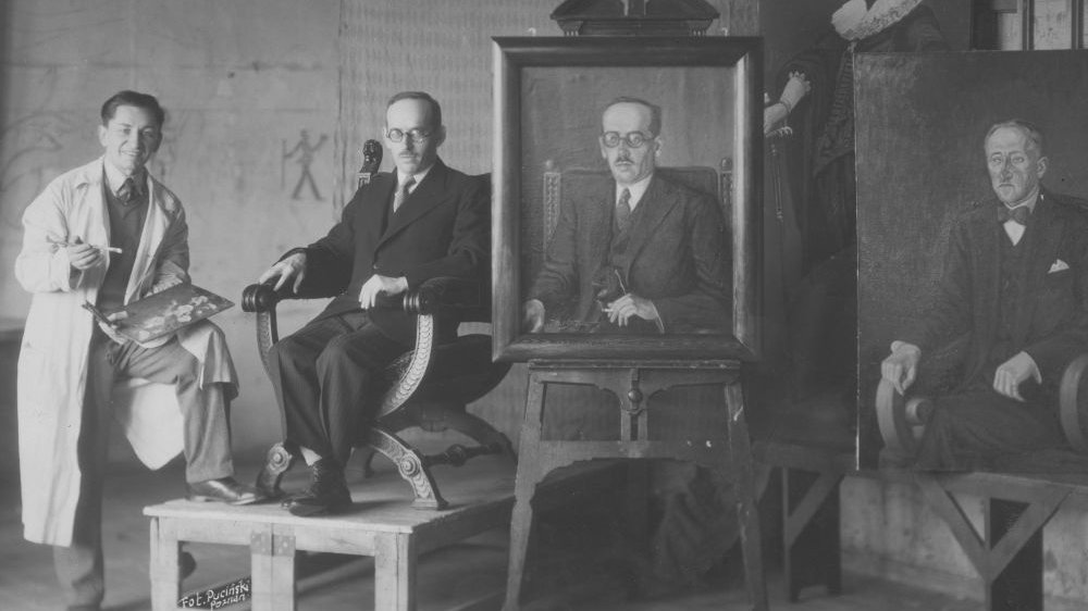 Mężczyzna w okularach i garniturze siedzi na ozdobnym fotelu na podwyższeniu, obok niego stoi malarz w białym fartuchu i z paletą farb oraz pędzlem w dłoni. Po prawej znajduje się kilka portretów, w tym portret mężczyzny w okularach.