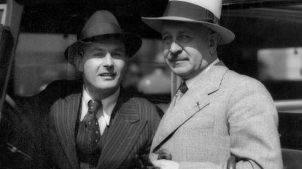 Dwaj eleganccy mężczyźni w garniturach i kapeluszach. Jeden siedzi w samochodzie, drugi stoi obok. Fotografia czarno-biała.