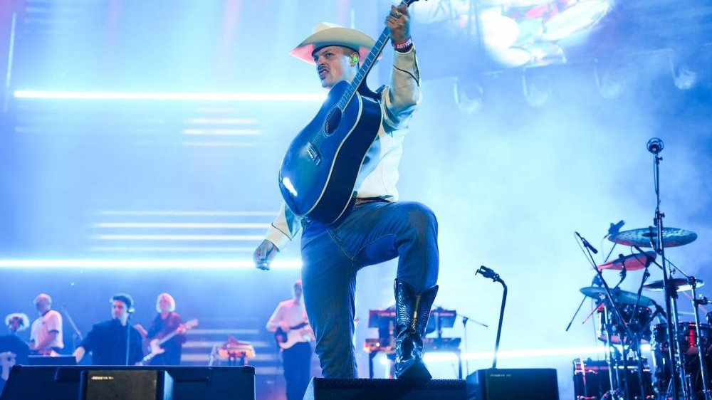 Mężczyzna w białym, kowbojskim kapeluszu, stoi na scenie z gitarą, jedną nogę stawia wyżej, na głośniku.