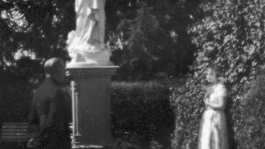 Filmowa scena spotkania kobiety i księdza w ogrodzie. Na środku kadry figura Matki Boskiej z dzieciątkiem Jezus na rękach.