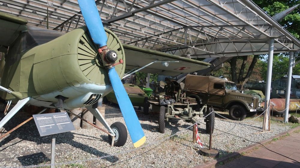 Pod zadaszeniem stoją eksponaty muzealne - stary samolot z niebieskim śmigłem oraz pojazd.