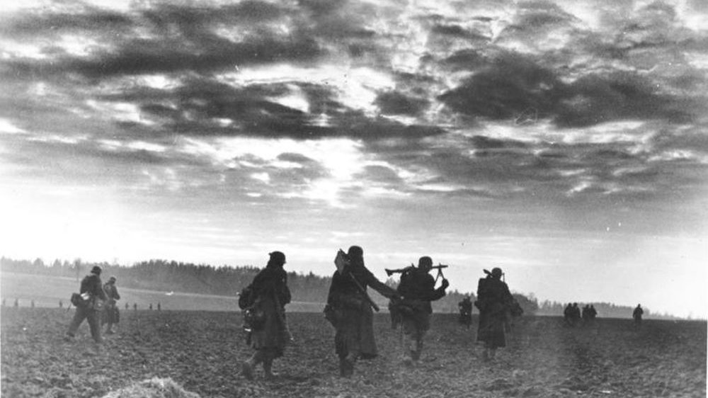 Czarno-biała fotografia idącej grupy żołnierzy z bronią. Nad nimi mocno zachmurzone niebo.