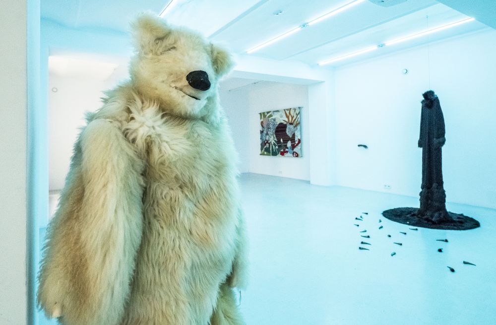 Biała sala wystawowa z kostiumem niedźwiedzia polarnego po lewej, czarnym kostiumem po prawej stronie i kolorową makatką w tle. - grafika artykułu