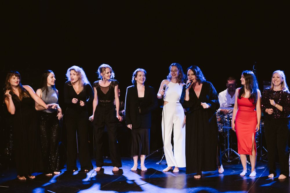 Na scenie stoi grupa kobiet w różnym wieku - wszystkie ubrane elegancko, dominują czarne kolory, ale dwie z nich mają na sobie białą i czerwoną kreację. Jedna z nich śpiewa do mikrofonu. - grafika artykułu