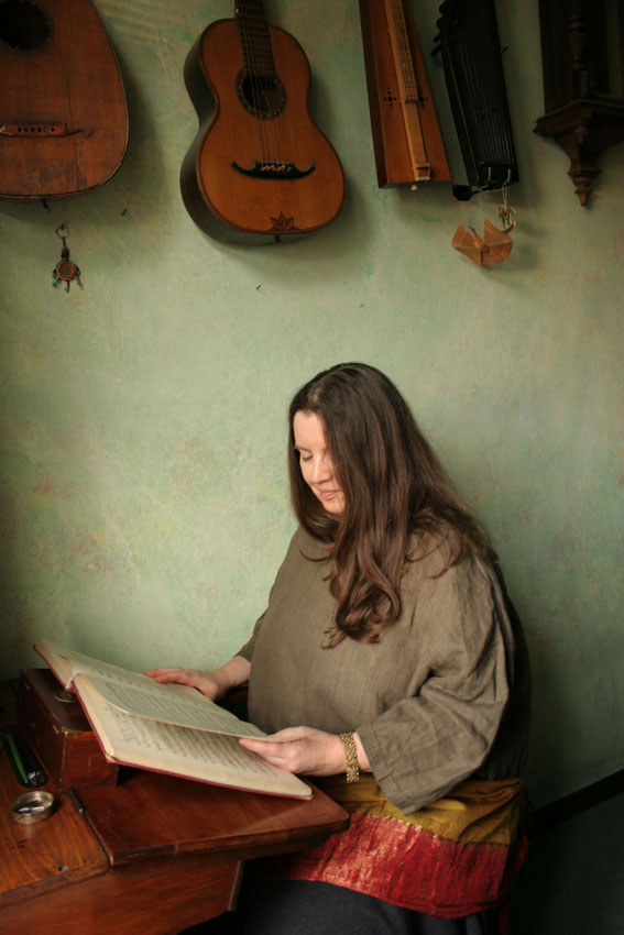 Artystka ma długie brązowe włosy, siedzi przy drewnianym biurku i przegląda dużą książkę, nad nią, na ścianie, wiszą instrumenty smyczkowe. - grafika artykułu