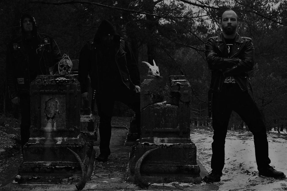 Ciemna fotografia w czarno-białych kolorach, trzech ubranych na czarno mężczyzn stoi w otoczeniu nagrobków, na których spoczywają przedmioty przypominające zwierzęce czaszki. - grafika artykułu