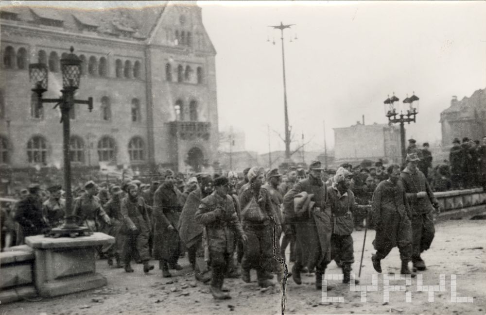 Czarno-biała fotografia idącej grupy mężczyzn. Za ich plecami budynek Zamku Cesarskiego. - grafika artykułu
