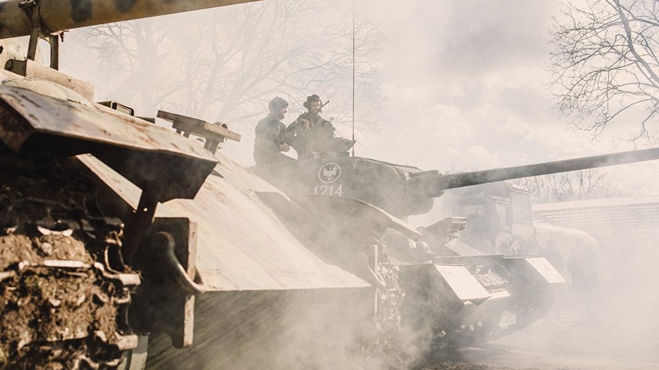 Czołg T-34 z polskimi oznaczeniami na wieży owiany dymem spalin, na wieży czołgiści, obok inny czołg