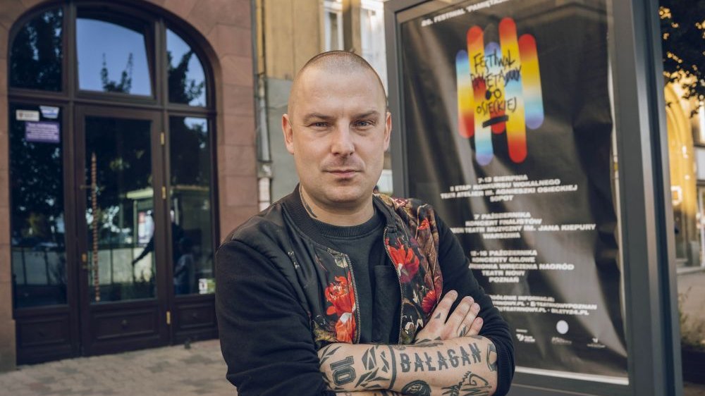 Artysta stoi obok plakatu reklamującego festiwal, przed wejściem do Teatru Nowego. Jest łysy, ma ramiona pokryte tatuażami oraz skórzaną kurtkę w czerwone wzory.