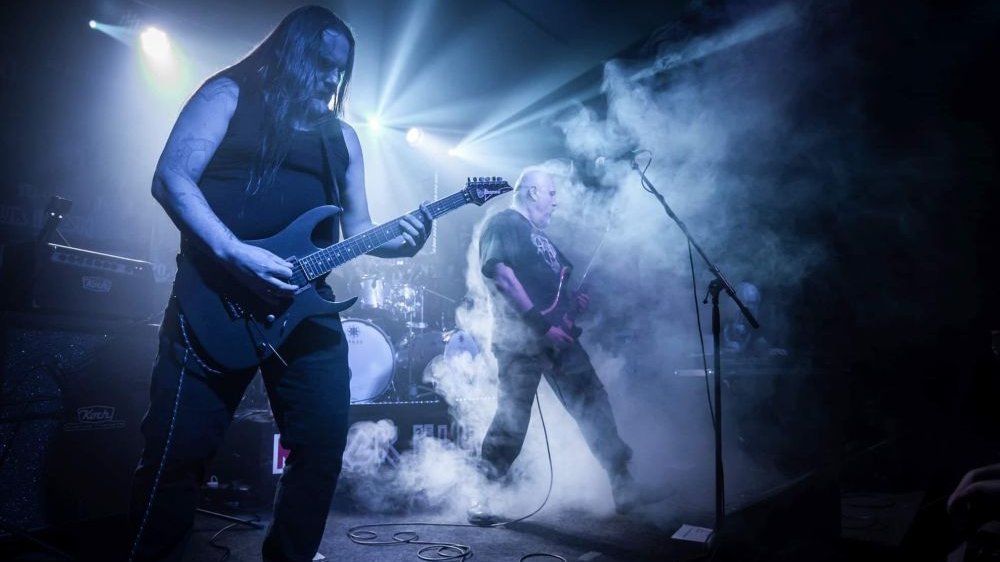 Zespół na scenie, na pierwszym planie długowłosy gitarzysta. Scena pokryta sztyczną mgłą, oświetlona na chłodny niebieski kolor.