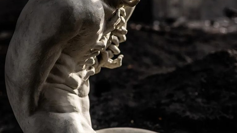 Rzeźba siedzącego korpusu ludzkiego, z którego szyi i piersi wystają fragmenty dłoni.