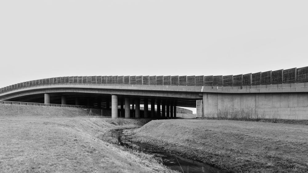 Płytka rzeczka nad którą ciągnie się wiadukt ogrodzony płotem/ścianami. Foto czarno-białe.