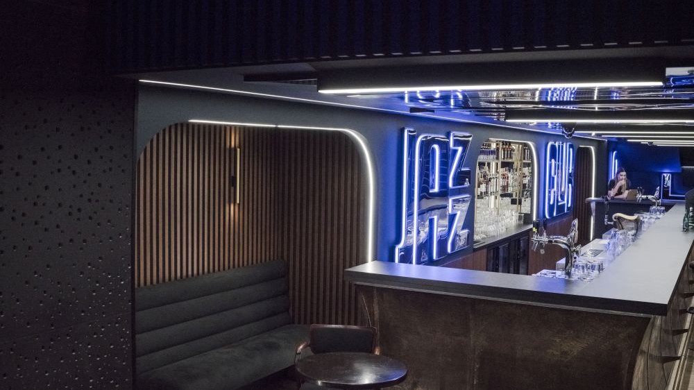 Fragment długiego baru z jednym krzesłem barowym. O boczną ścianę barową opiera się szara kanapa. Za barem kraniki, szklanki, kufle oraz jarzący się na niebiesko napis "Jazz Club".