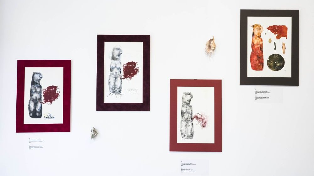 Cztery grafiki na białej ścianie galerii. Na grafikach postać kobieca i elementy towarzyszące, między innymi czerwony kłębek nici.