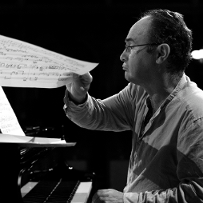 Black and white photo of a man sitting at the piano. He holds paperpage with music notes in his hand.