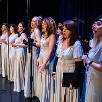 Photo from the concert: women in long white-silver dresses singing on stage.