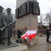 Photo of the monument to the Greater Poland Insurgents. A man speaking in front of the monument and white and red Polish flags, soldiers around the monument.