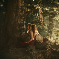 Two girls in dresses are sitting under a large tree, leaning against each other, each holding an old-fashioned drum in her arms. One of them is illuminated by the sun.