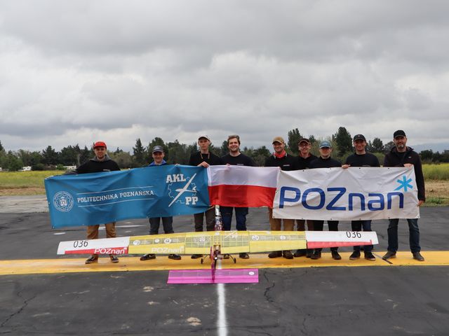 A group of young men standing on the runway behind a lying model airplane. The men hold the banner of the Poznań University of Technology, the Polish flag and the banner of the City of Poznań. - grafika artykułu